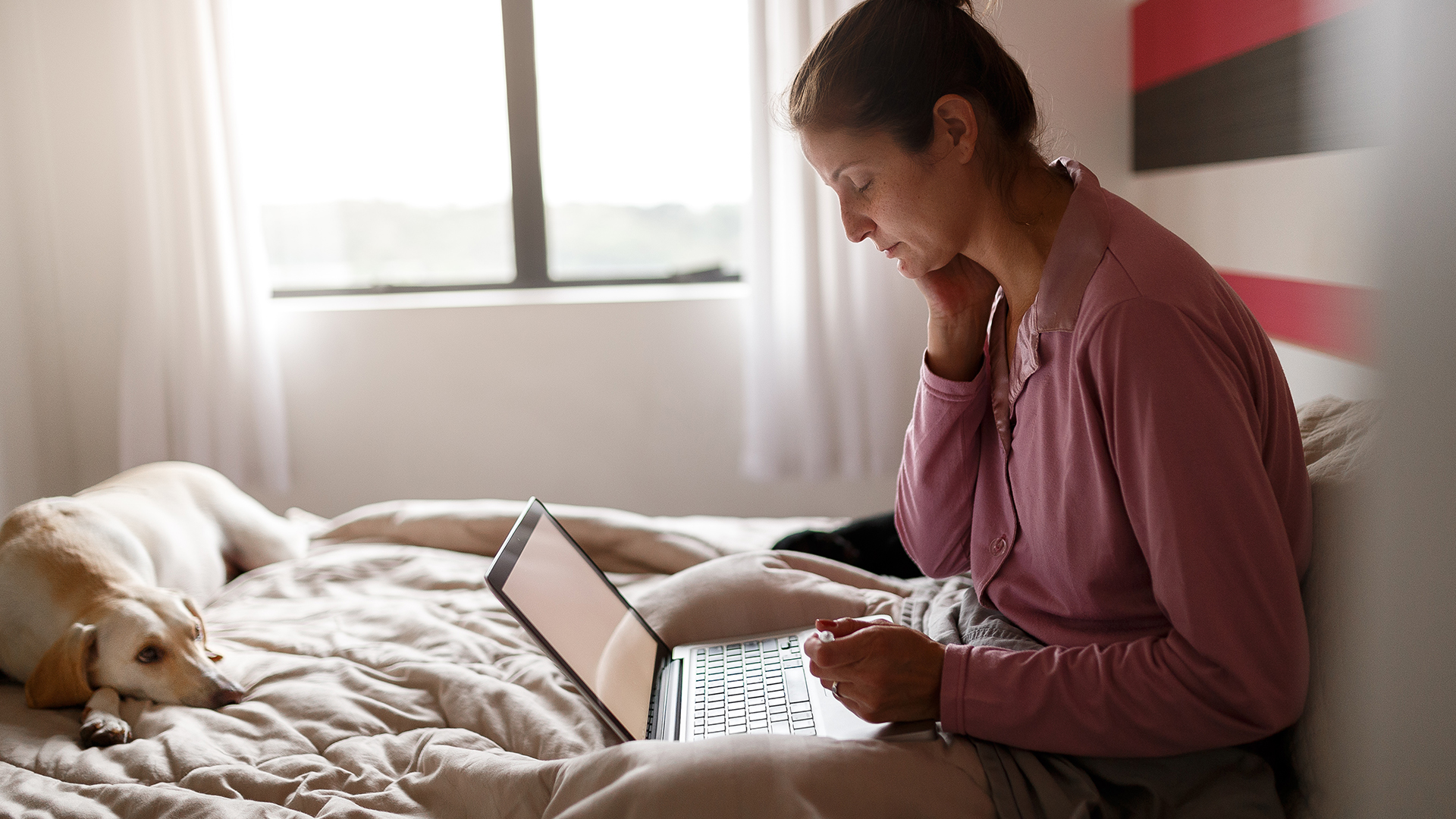 A middle aged woman looks at her laptop pensively from the comfort of her bed while her dog looks on.